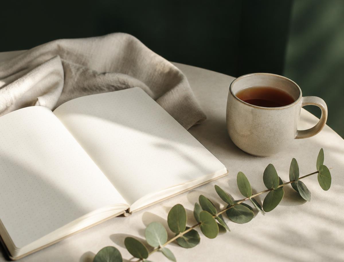 An open notebook, a ceramic mug of tea and a sprig of eucalyptus on a linen-covered table